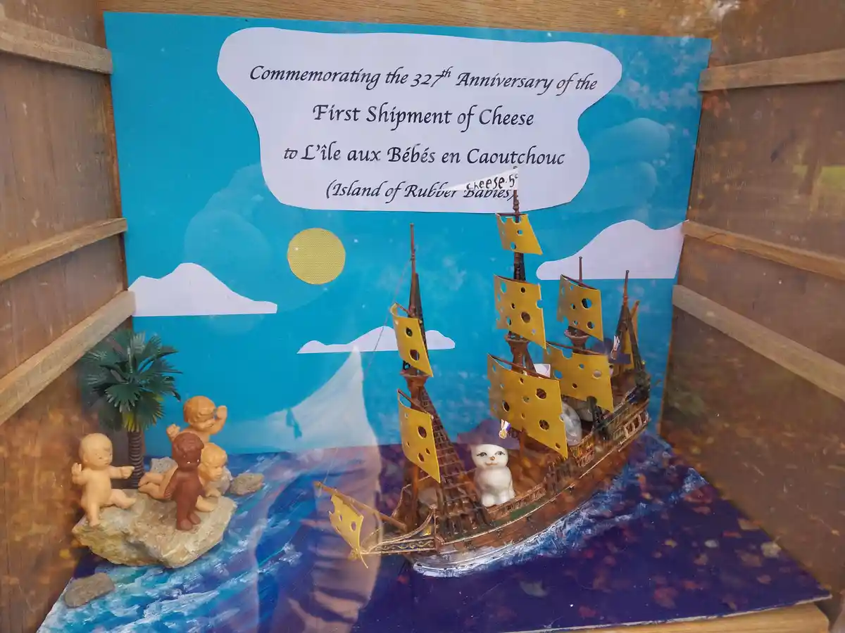 A diorama depicts a toy ship with cheese-patterned sails sailing toward a shore with rubber baby figurines, under a blue sky…