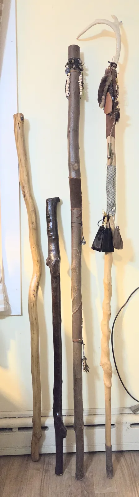 Five rustic walking sticks of varying shapes and sizes are leaning against a pale wall on a wooden floor. The sticks range…