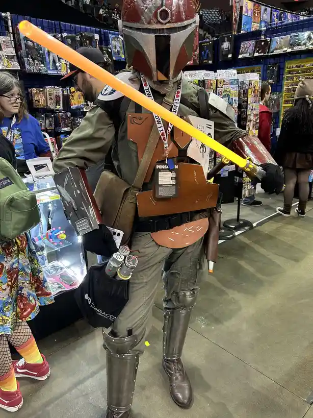 A person in a Mandalorian-style costume at a convention holds a bright orange lightsaber, with the text 'i'm not a jedi, i'm…