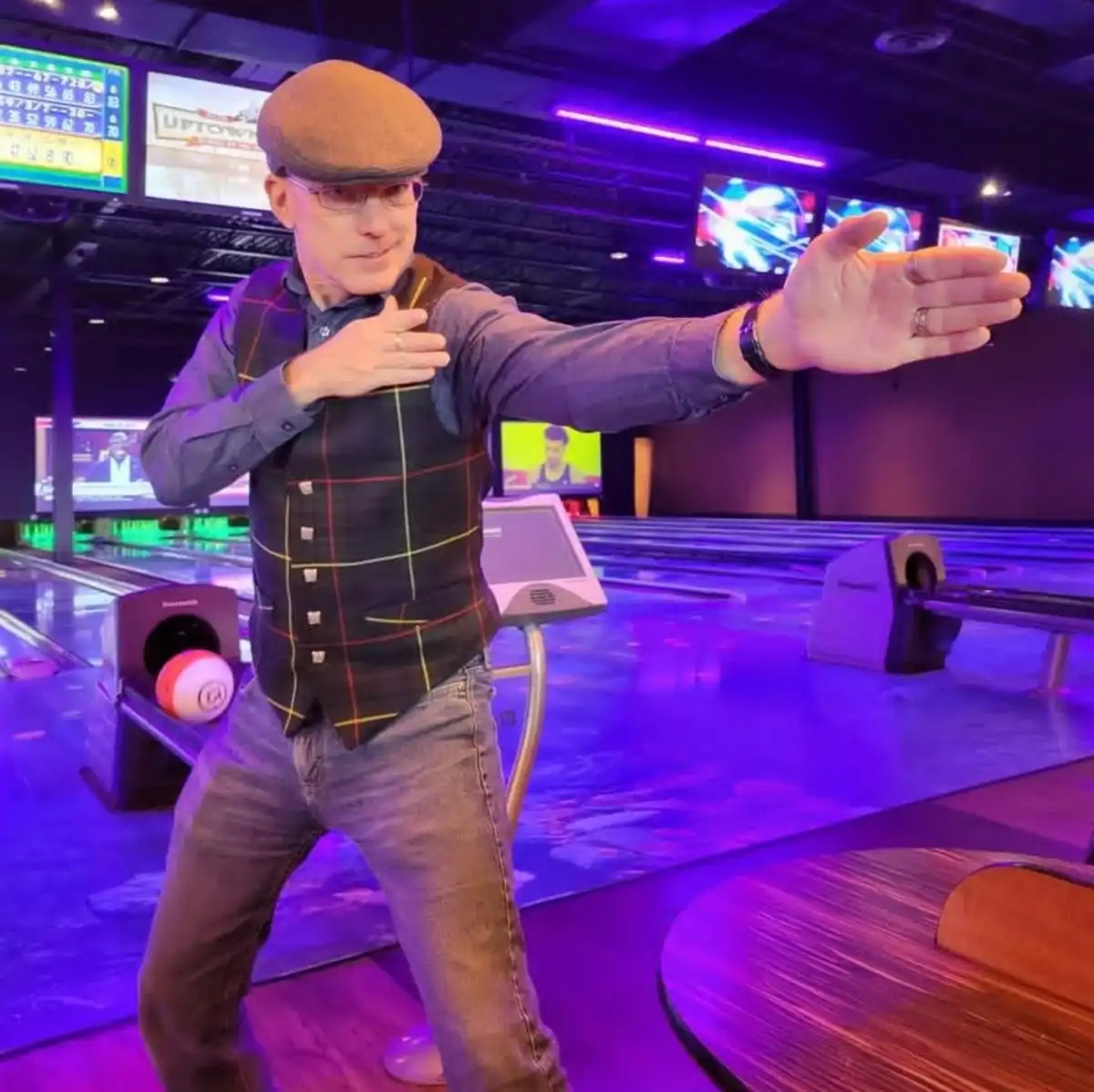 A man in a brown flat cap, glasses, and a plaid vest stands in a bowling alley with purple lighting, striking a pose with…