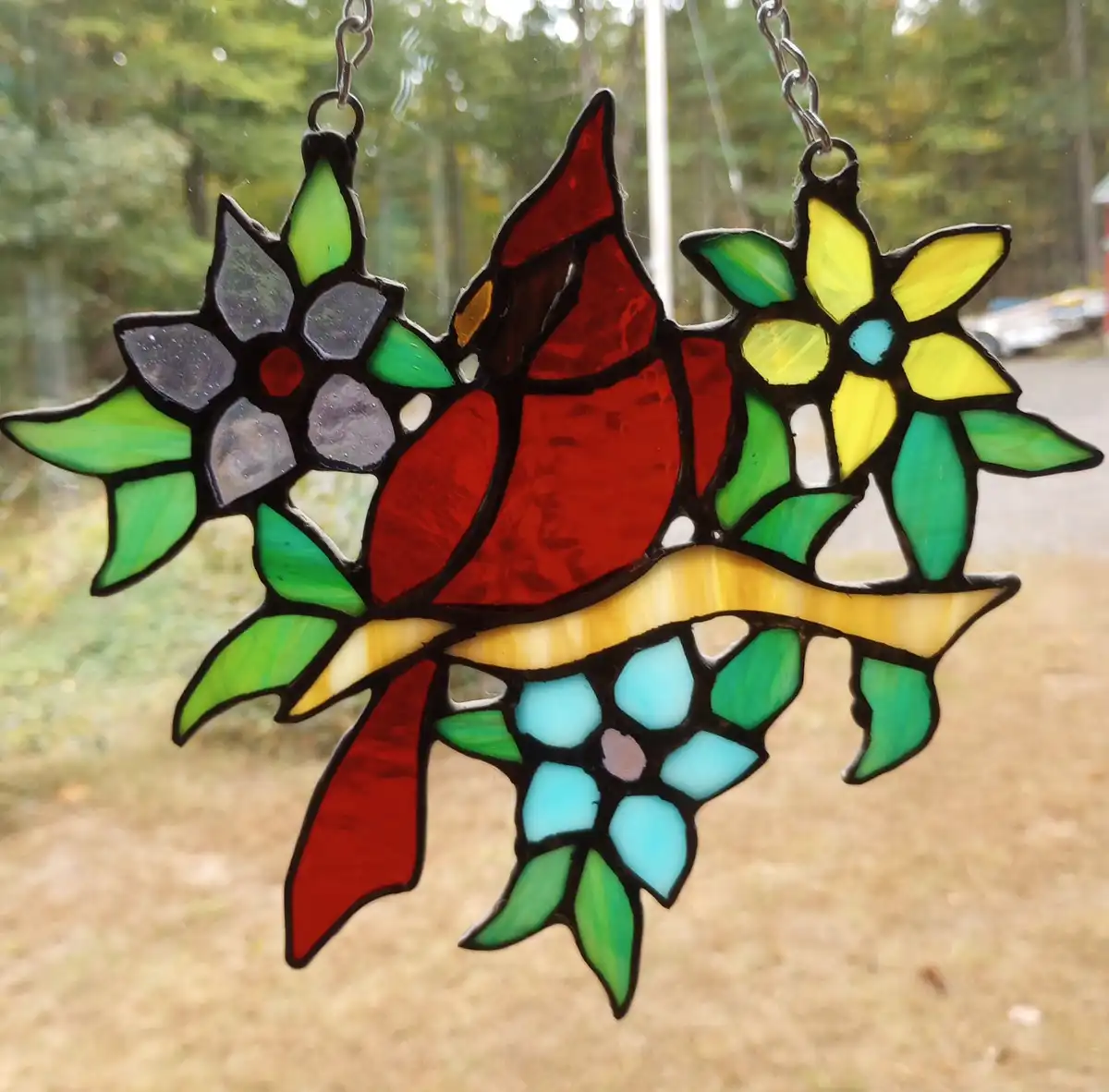 A stained glass window hanging depicts a red cardinal perched on a branch, flanked by colorful flowers and green leaves…