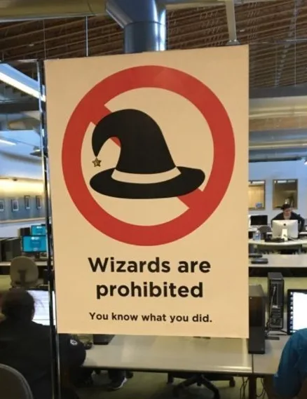 A sign in an office setting displays a red circle with a slash over a black wizard's hat icon. Below the icon, the text…