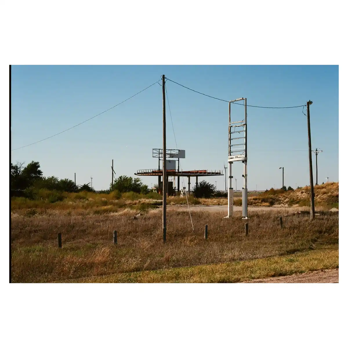 An abandoned, rusted gas station with a skeletal canopy and a tall, empty sign frame stands in a dry, grassy field under a…