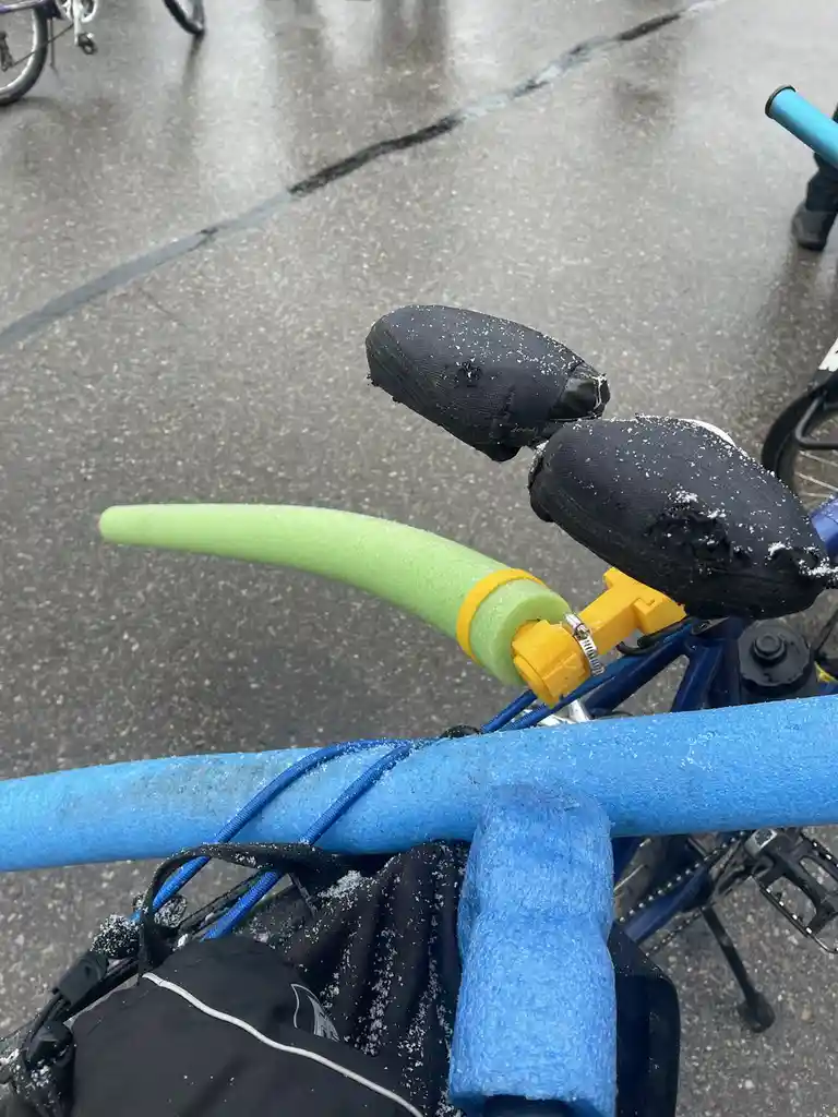 A close-up photo of a bicycle handlebar and stem covered in snow, with a green pool noodle wrapped around the grip and a…