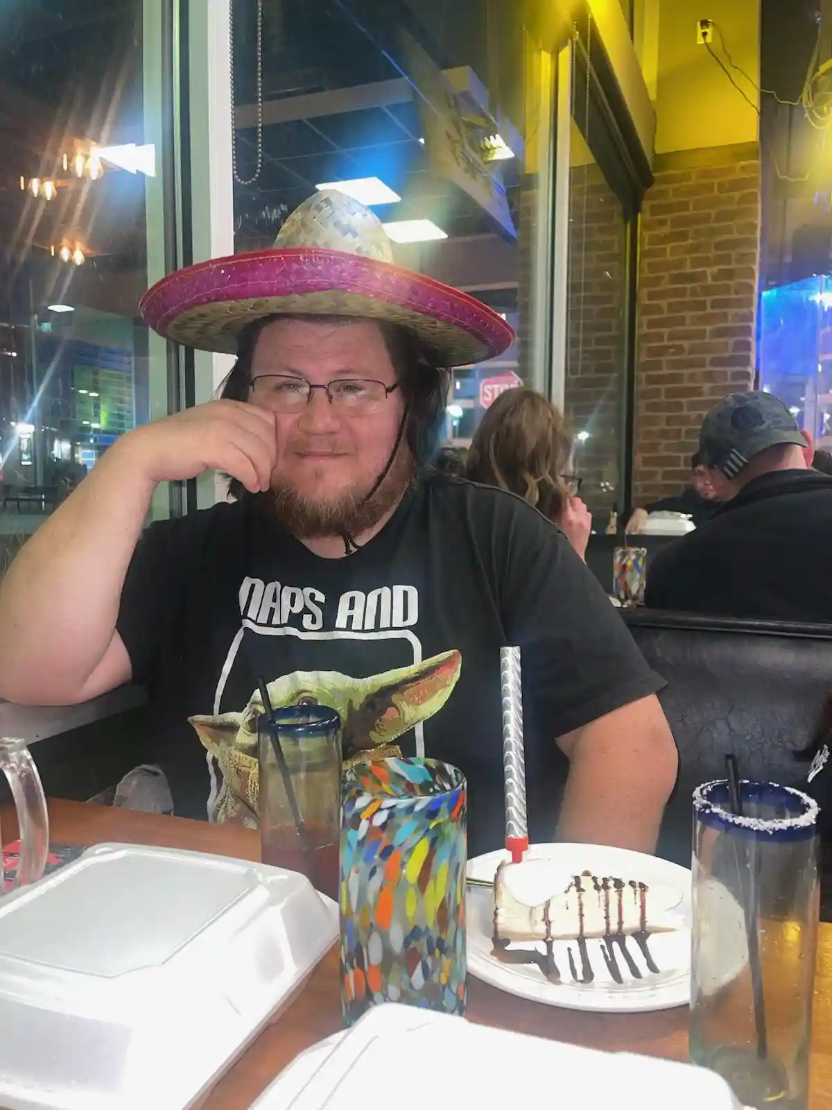 A man with glasses and a beard sits at a restaurant table wearing a large sombrero with a pink trim. He rests his chin on…