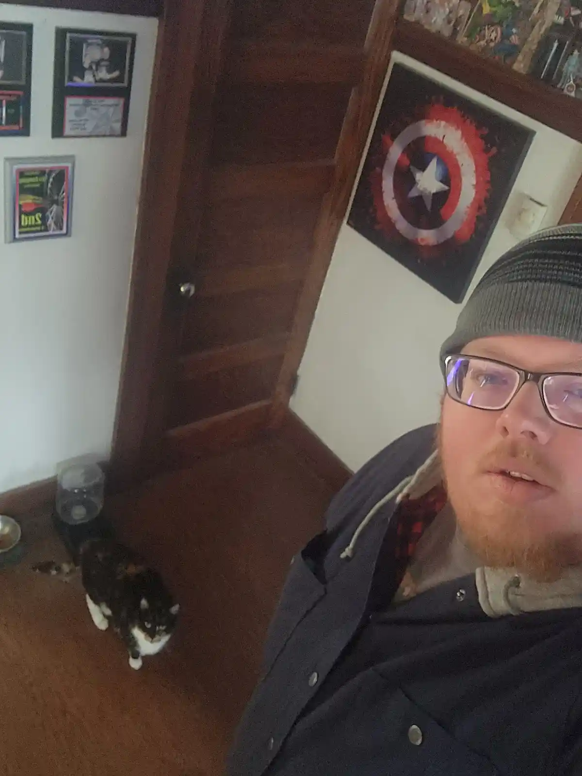 A man with glasses and a beanie takes a selfie in a room with a wooden door and walls decorated with framed pictures…