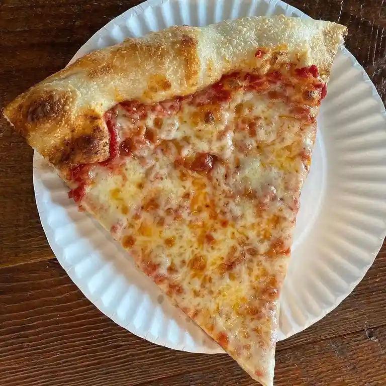 A single slice of cheese pizza with a golden-brown crust rests on a white paper plate atop a dark wooden table. The cheese…