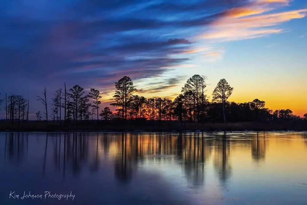 A serene landscape photo shows a frozen lake reflecting a vibrant sunset with silhouetted trees against a sky of orange…