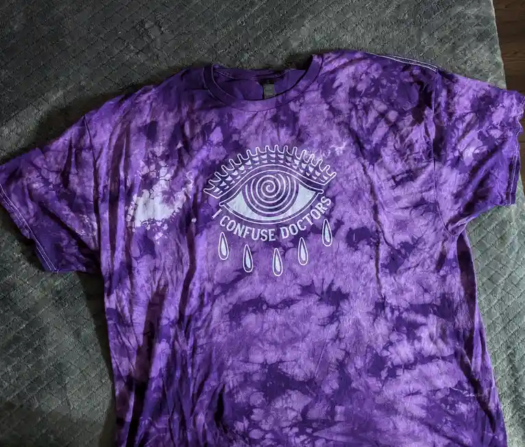 A purple tie-dye t-shirt with a white graphic of an eye with a spiral pupil and a crown-like top, surrounded by the text 'I…