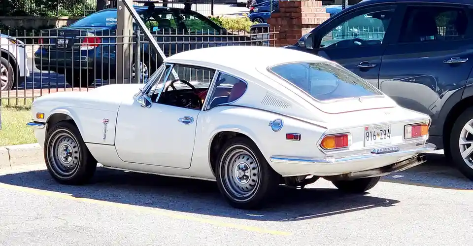 A white vintage Triumph TR6 sports car is parked in a lot, featuring a Maryland license plate that reads '816 Z84' with…