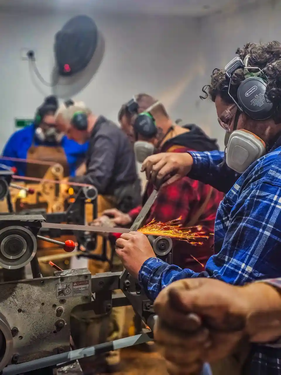 Several people wearing safety goggles and ear protection are using belt grinders in a workshop, with sparks flying as they…