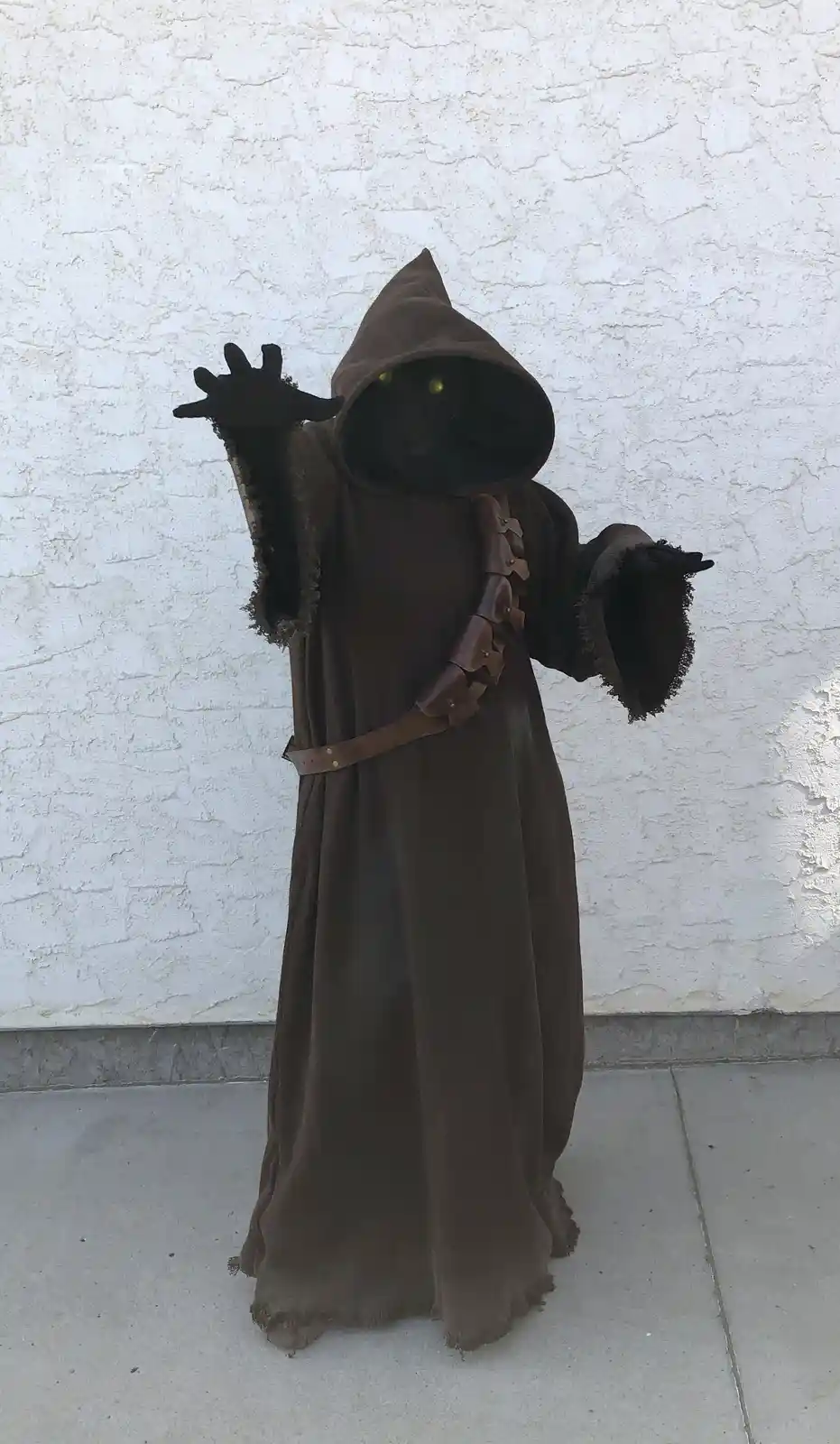 A person in a full-body brown hooded robe with glowing yellow eyes and black gloves stands against a white stucco wall, one…
