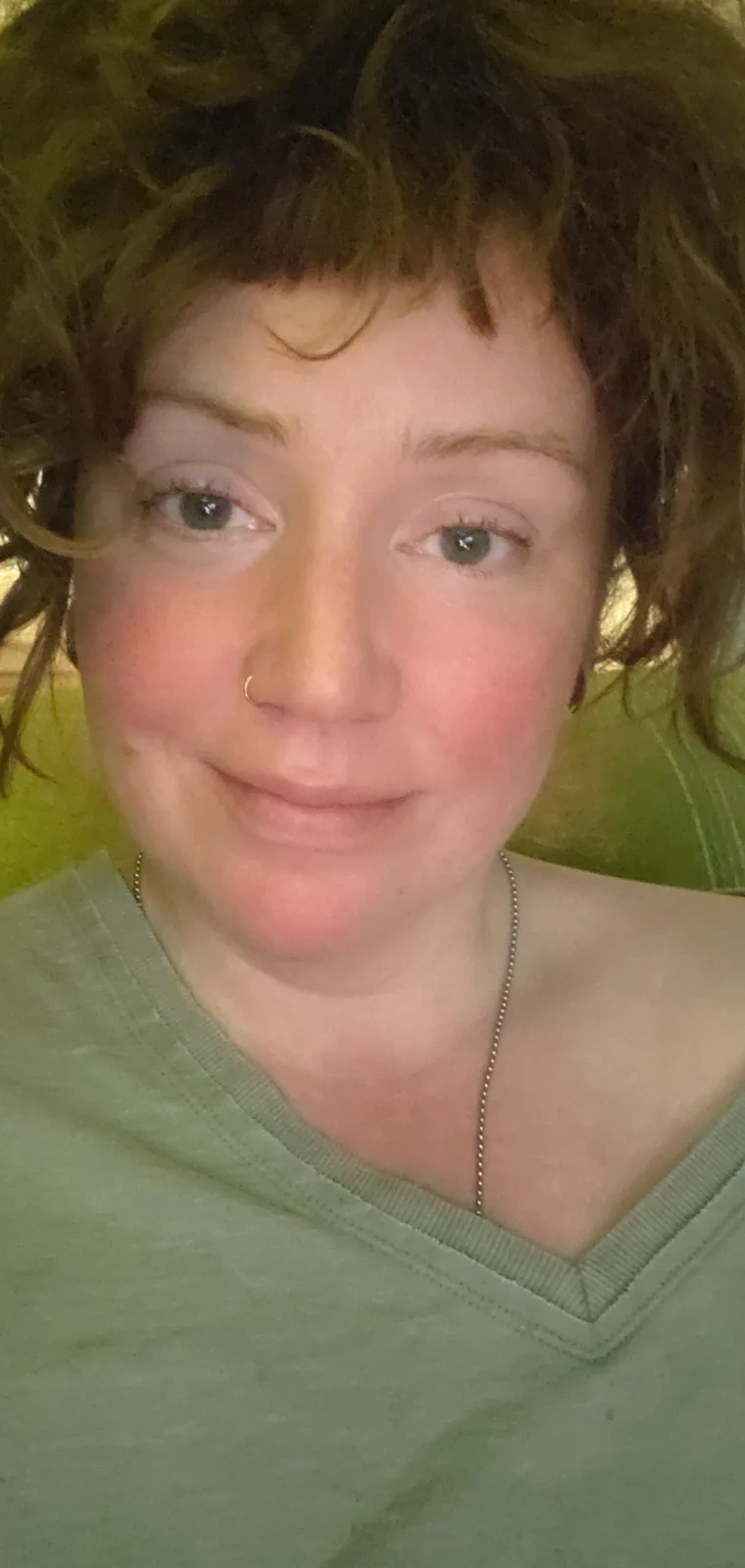 A close-up selfie of a person with short, curly brown hair, wearing a green v-neck shirt and a thin chain necklace, with a…