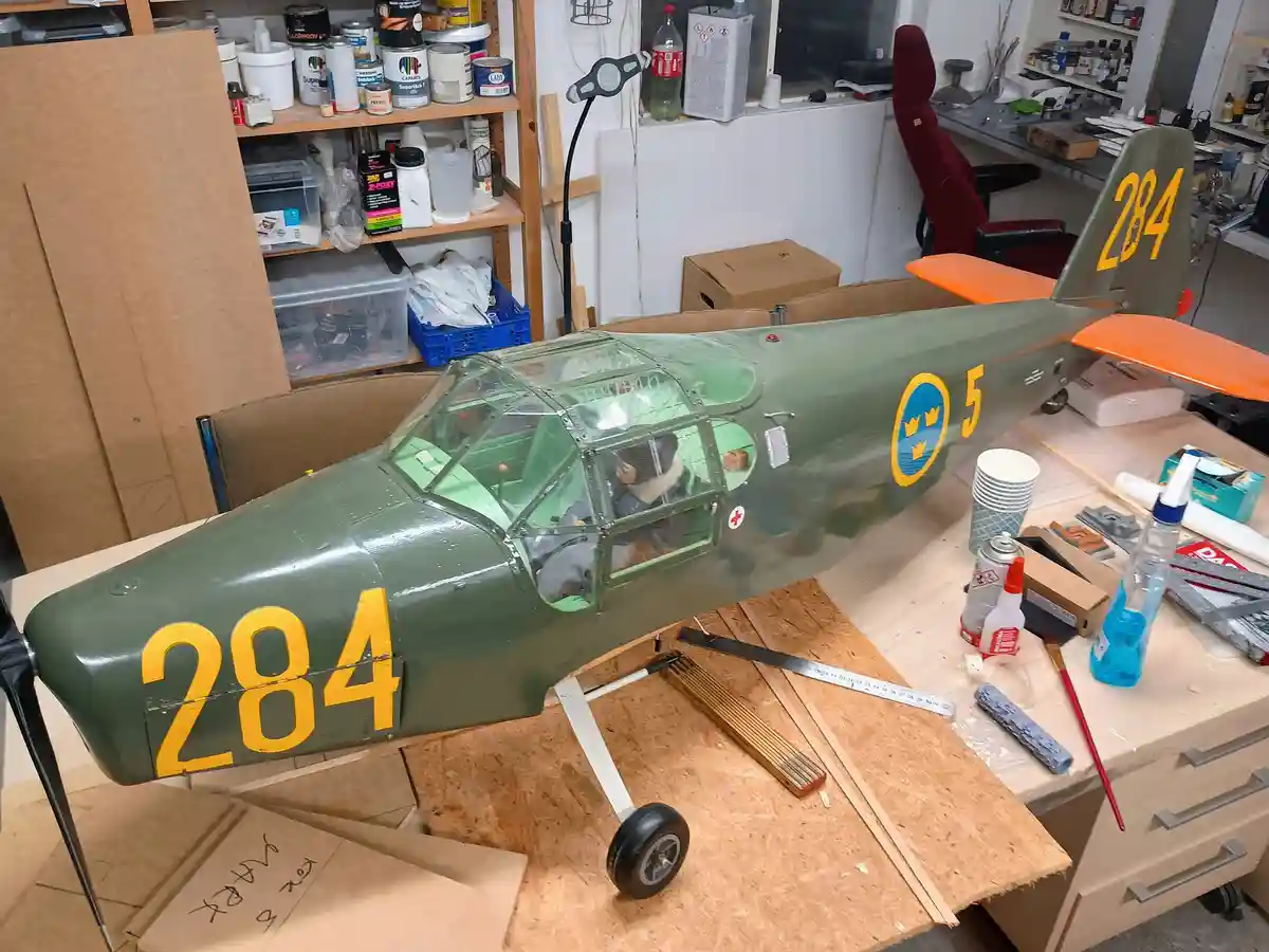 A large, green model airplane with yellow markings '284' on the nose and tail sits on a workbench in a cluttered hobbyist…