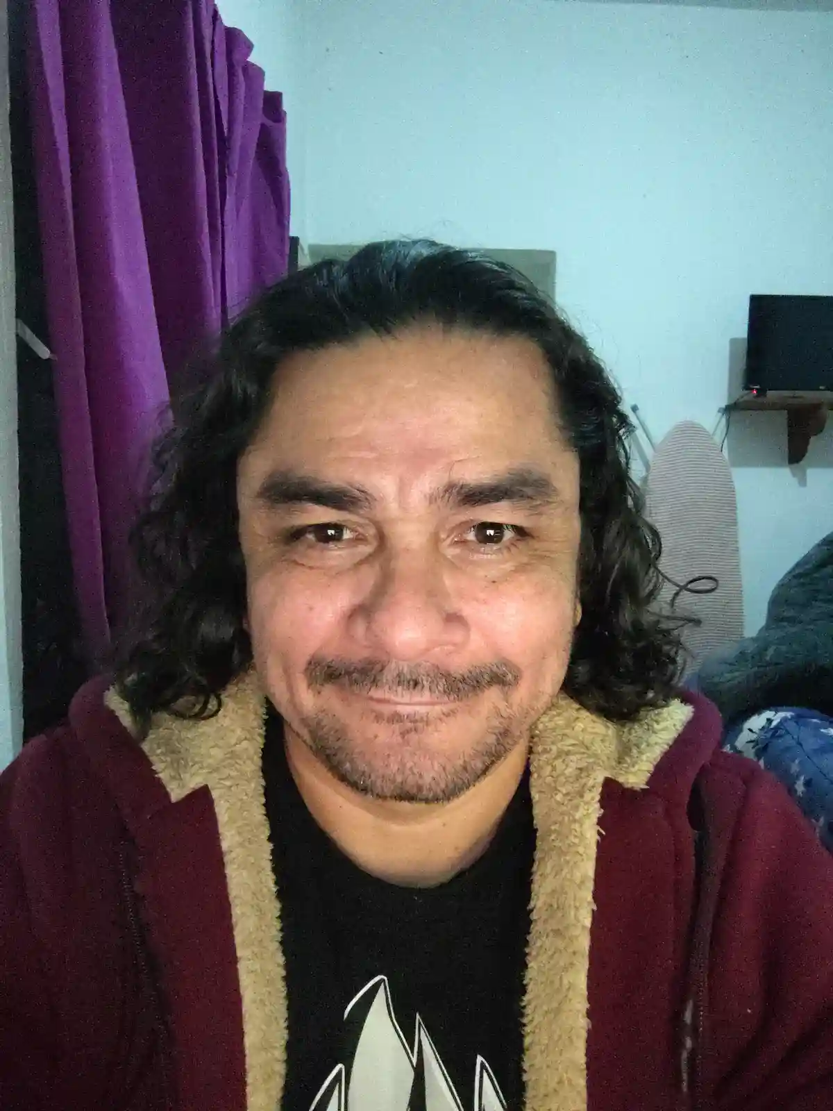 A man with long dark curly hair and a goatee smiles slightly while wearing a maroon fleece-lined hoodie over a black…