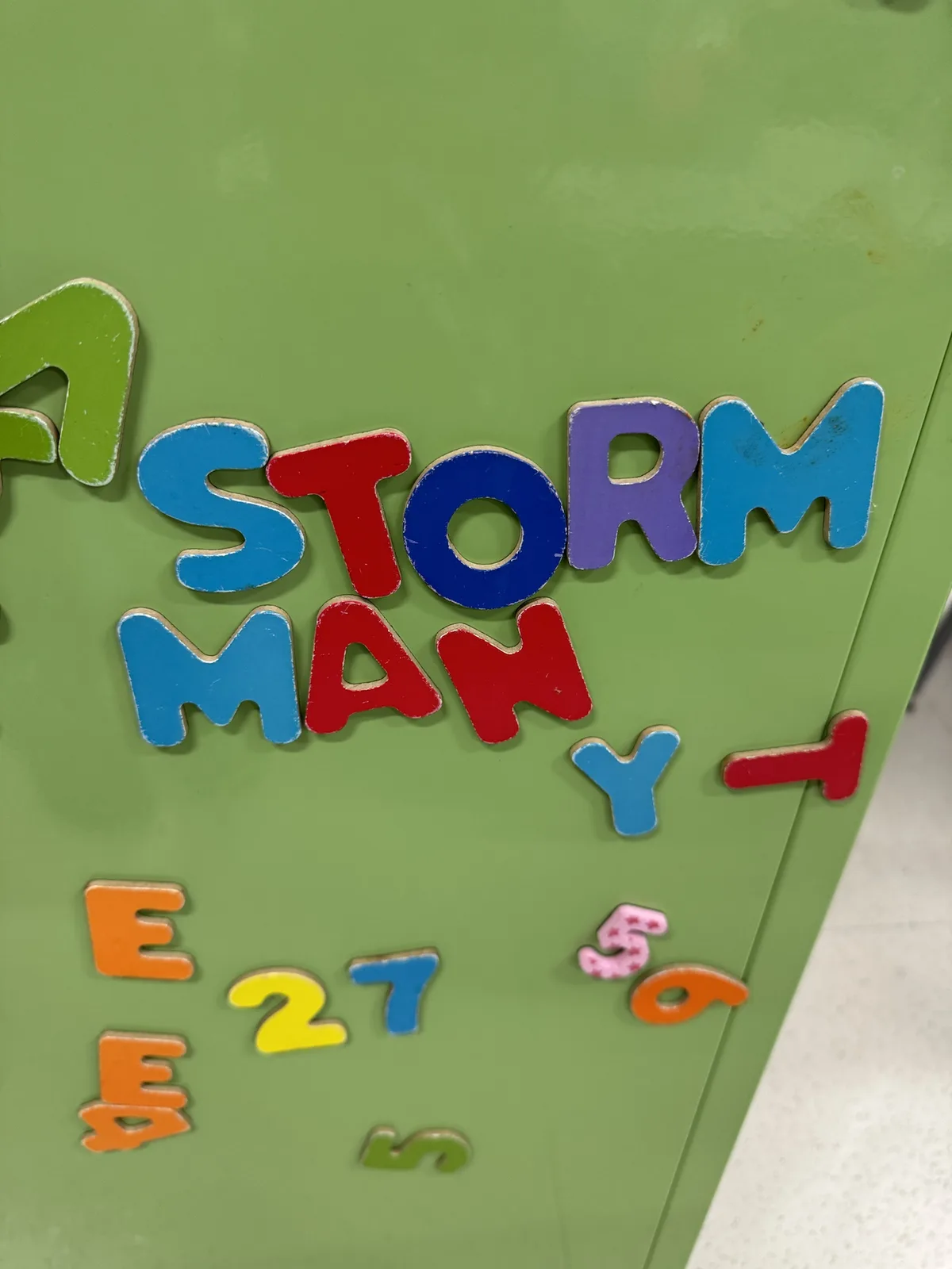 A close-up of colorful magnetic letters and numbers stuck to a green surface, forming the phrase 'A STORM MAN Y-' with the…