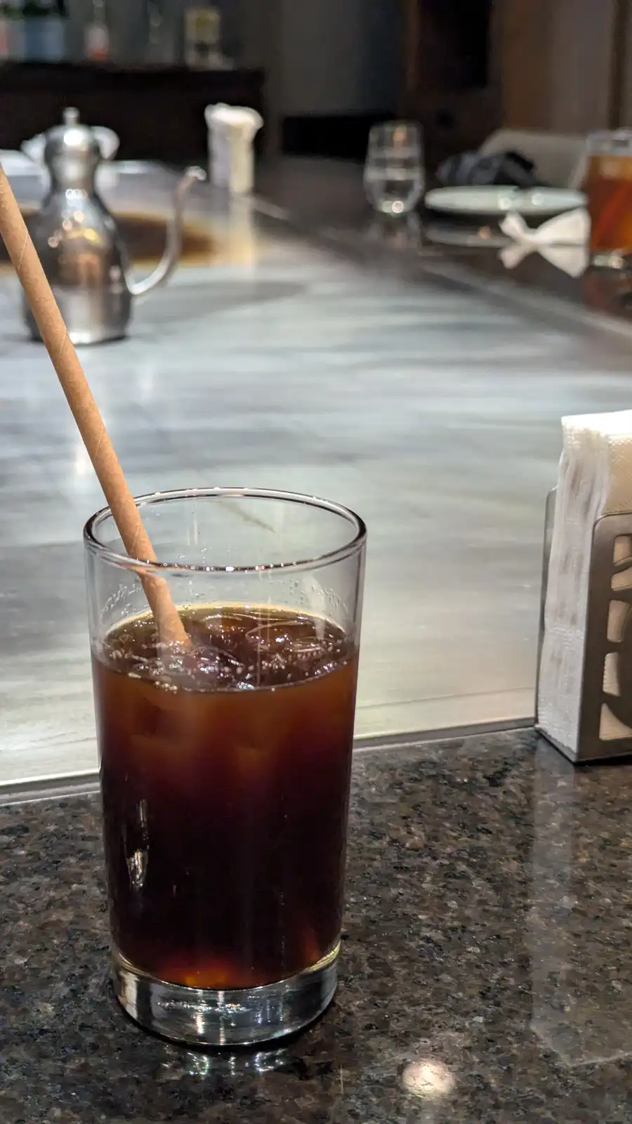 A glass of iced coffee with a wooden stirrer sits on a dark granite bar counter, with a blurred background showing a…