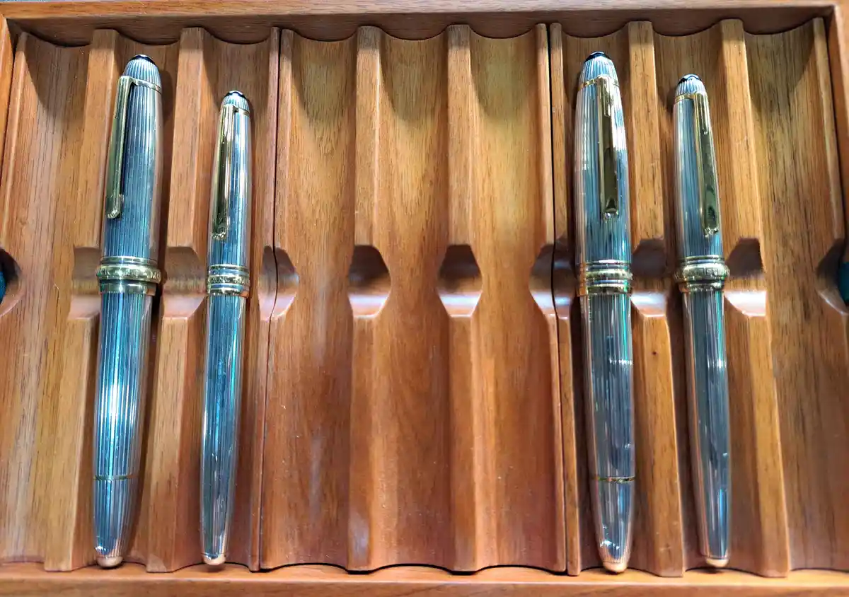 Four elegant, silver-and-gold pens are neatly arranged in a wooden display case with carved slots. Two pens are on the left…