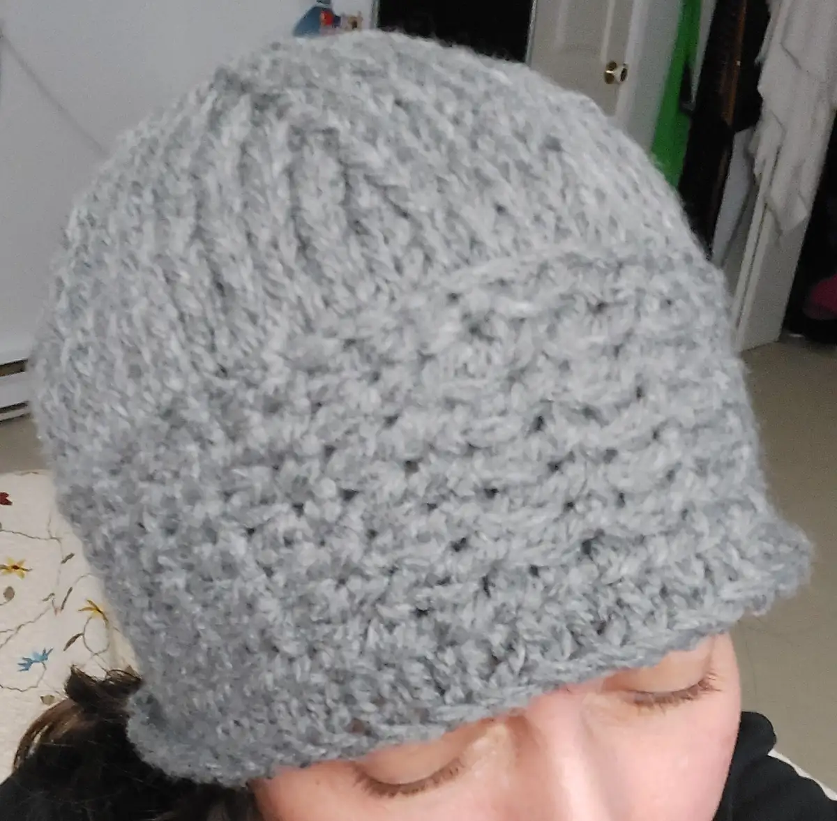A close-up photo shows a person wearing a light gray, textured crocheted beanie that covers their eyes, with only their…
