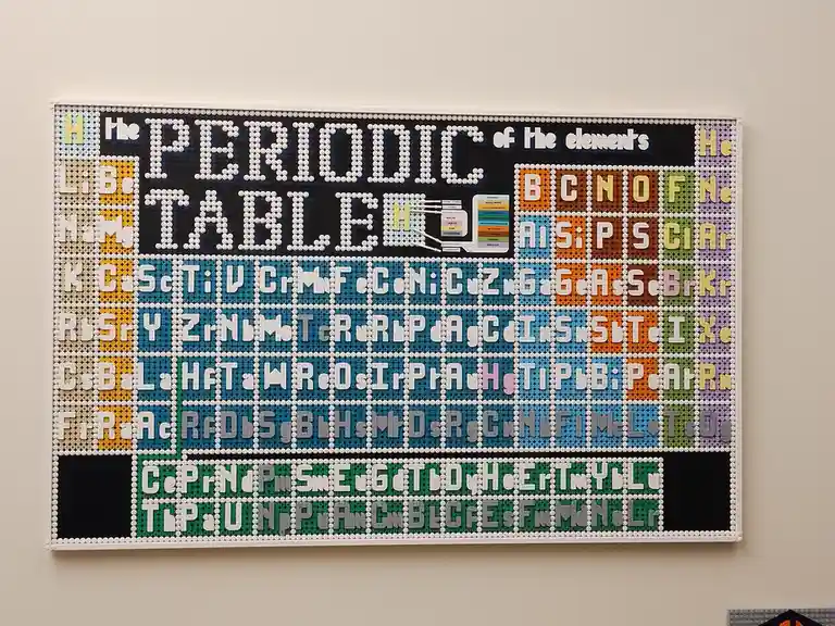 A large, wall-mounted periodic table constructed from colorful LEGO bricks, displaying element symbols in white lettering on…