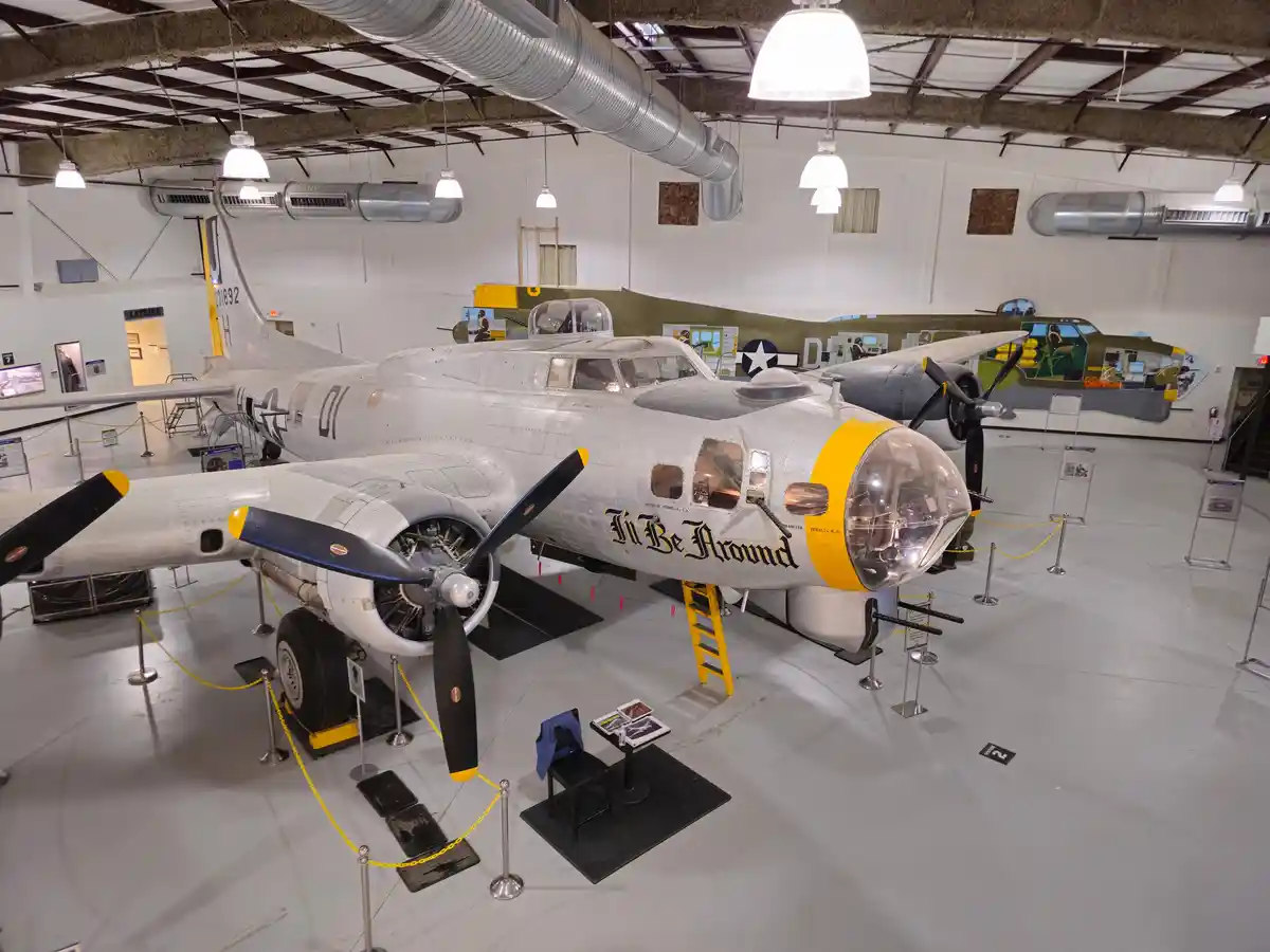 A silver WWII-era bomber aircraft, likely a B-17 Flying Fortress, is displayed indoors in a museum with yellow-tipped…