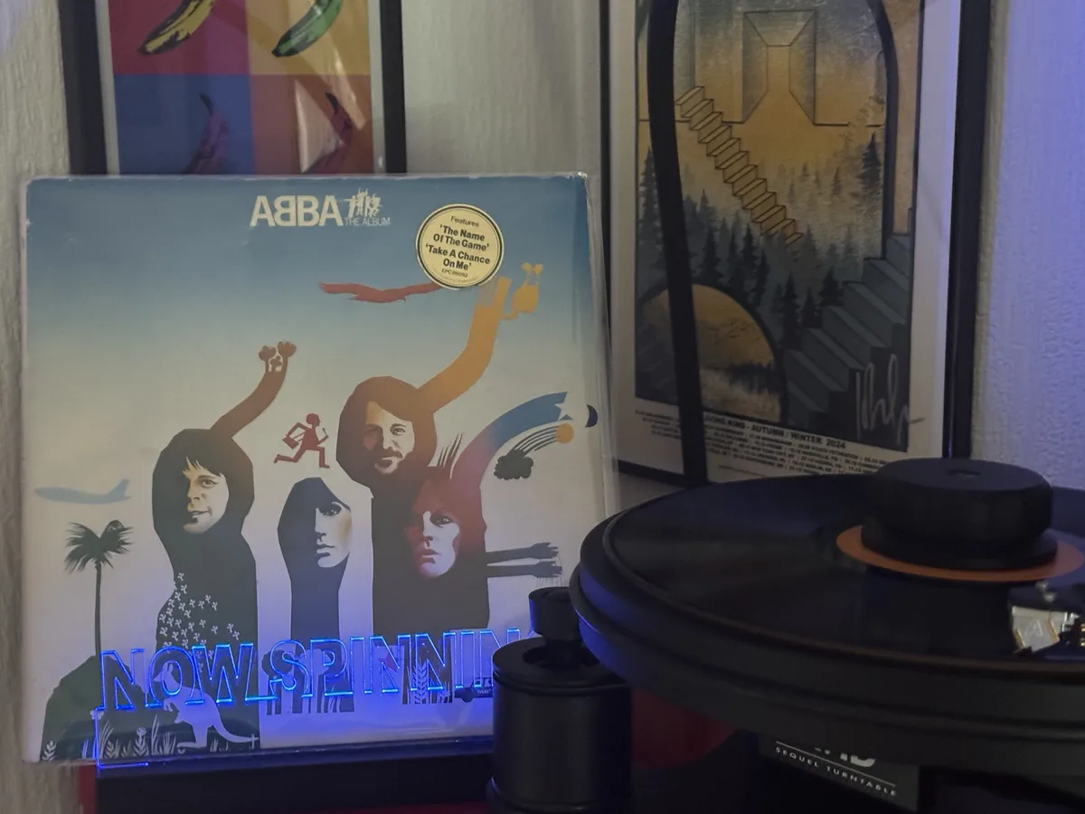 A vinyl record player with a record on it sits next to the album cover for ABBA's 'The Album'. The cover art features…