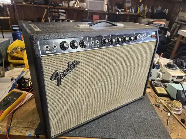 A vintage Fender Deluxe Reverb guitar amplifier sits on a cluttered workbench surrounded by tools, soldering equipment, and…