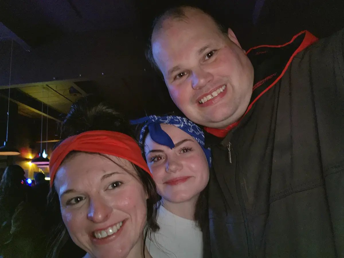 Three people pose closely for a selfie in a dimly lit indoor setting, likely a bar or club, with hanging lights visible in…