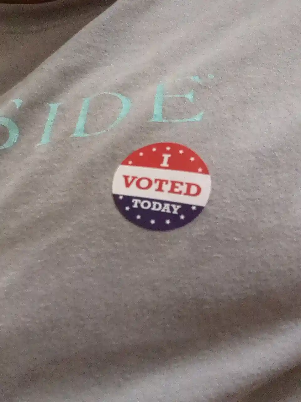 A close-up of a gray sweatshirt with light blue embroidered text partially visible as 'SIDE'. Affixed to the shirt is a…