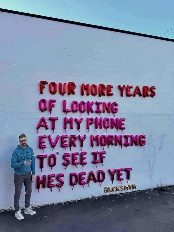 A man in a blue hoodie and glasses stands beside a white wall, looking at his phone. The wall features large, pink…