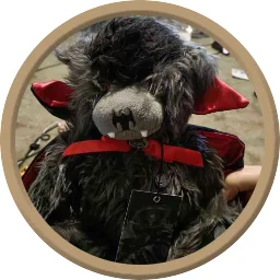 A close-up photo of a black, furry plush toy resembling a devil or demon with red horns, red cape, and fangs, wearing a red…