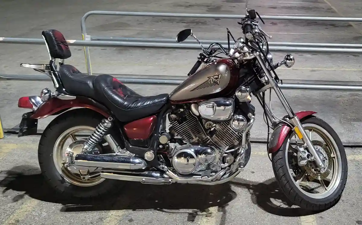 A red and chrome Yamaha Virago 1997 motorcycle is parked at night in a paved lot with metal railings in the background. The…