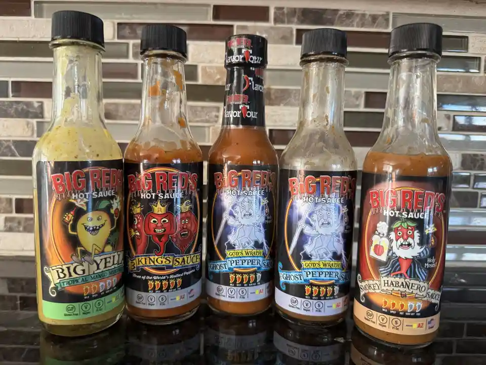 Five partially empty bottles of Big Red's Hot Sauce are lined up on a dark countertop against a tiled backsplash. Each…
