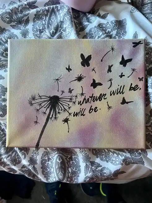 A canvas painting shows a black silhouette of a dandelion with seeds blowing away, some shaped like butterflies, against a…