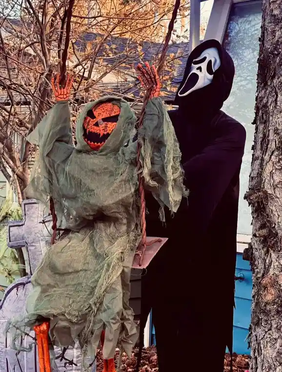 A person in a black Ghostface costume from 'Scream' stands beside a Halloween decoration of a pumpkin-headed ghost on a…