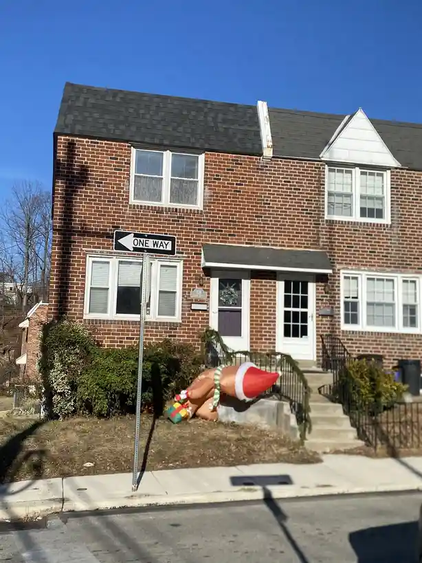 A brick house with a 'ONE WAY' street sign pointing left, next to a fallen inflatable Santa Claus decoration lying on the…