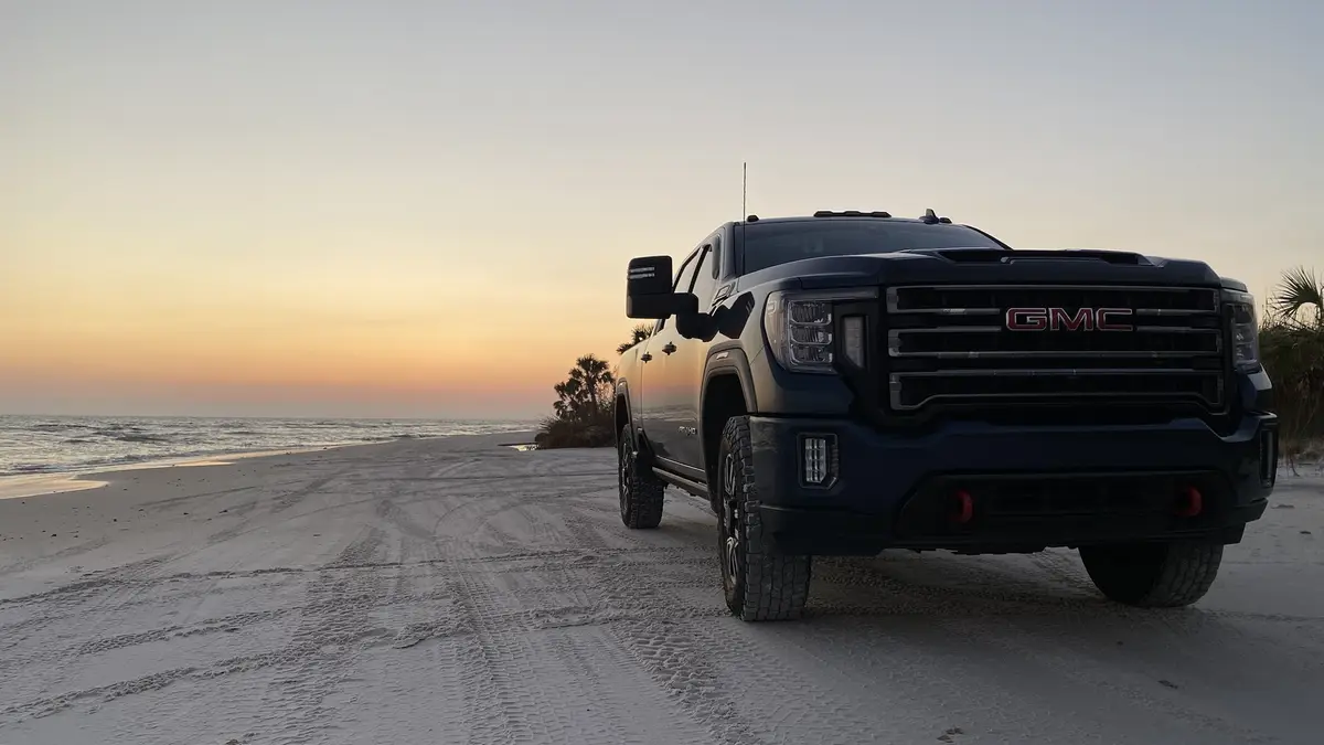 A dark blue GMC pickup truck is parked on a sandy beach with tire tracks in the sand, facing the camera. The ocean and a…