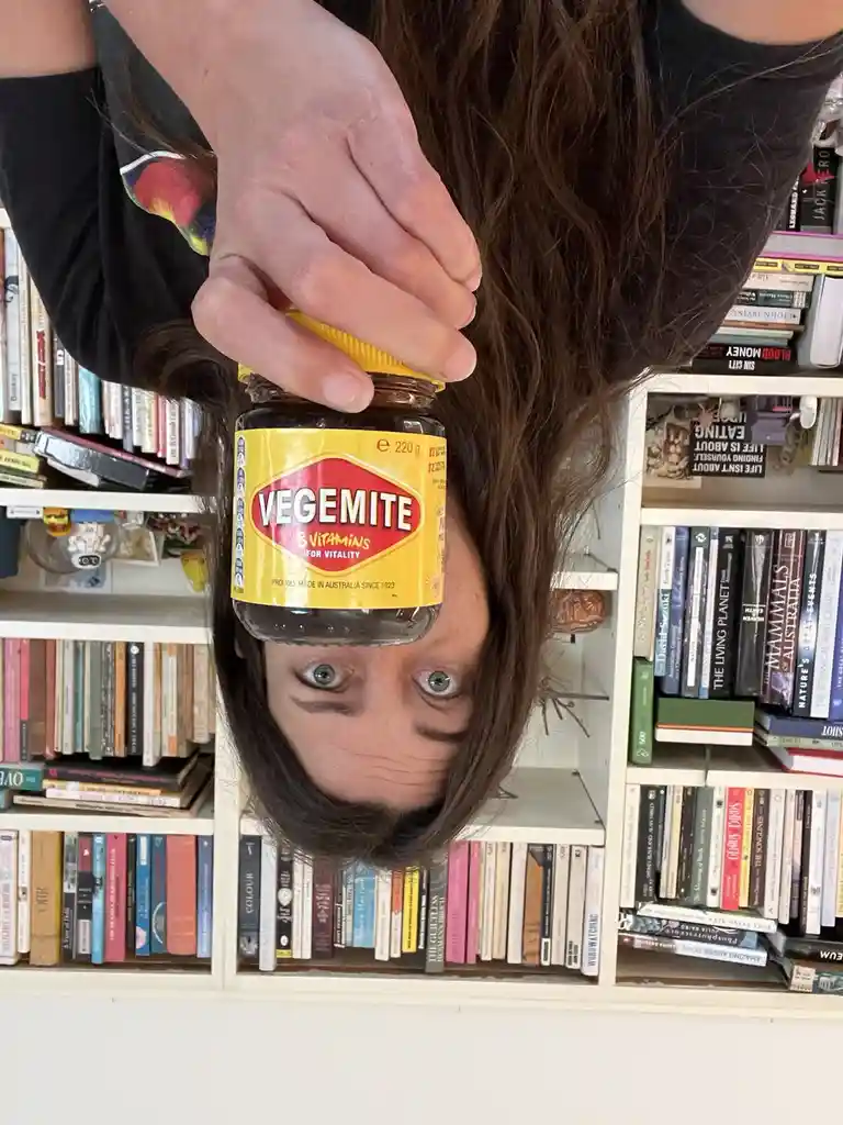 A person with long brown hair is upside down, holding a jar of Vegemite directly over their face, obscuring their nose and…