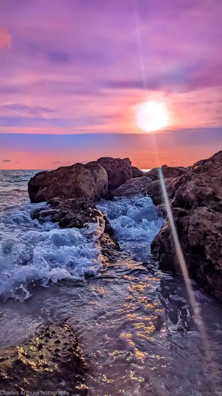 A serene sunset photograph shows waves crashing against dark rocks, with the sun low on the horizon casting pink and purple…