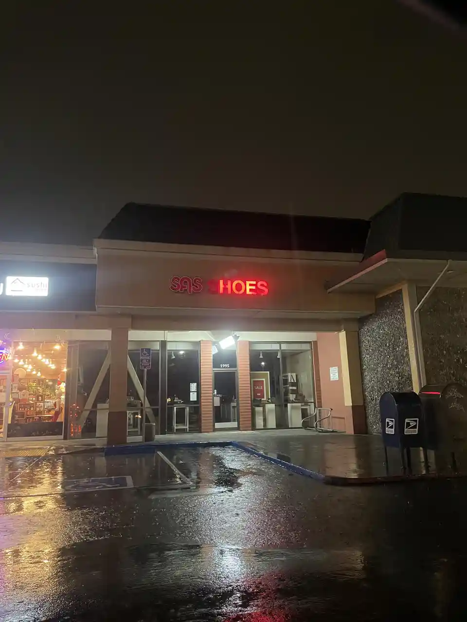 A nighttime photo of a wet parking lot in front of a store with a red neon sign that reads 'SAS SHOES', next to a sushi…