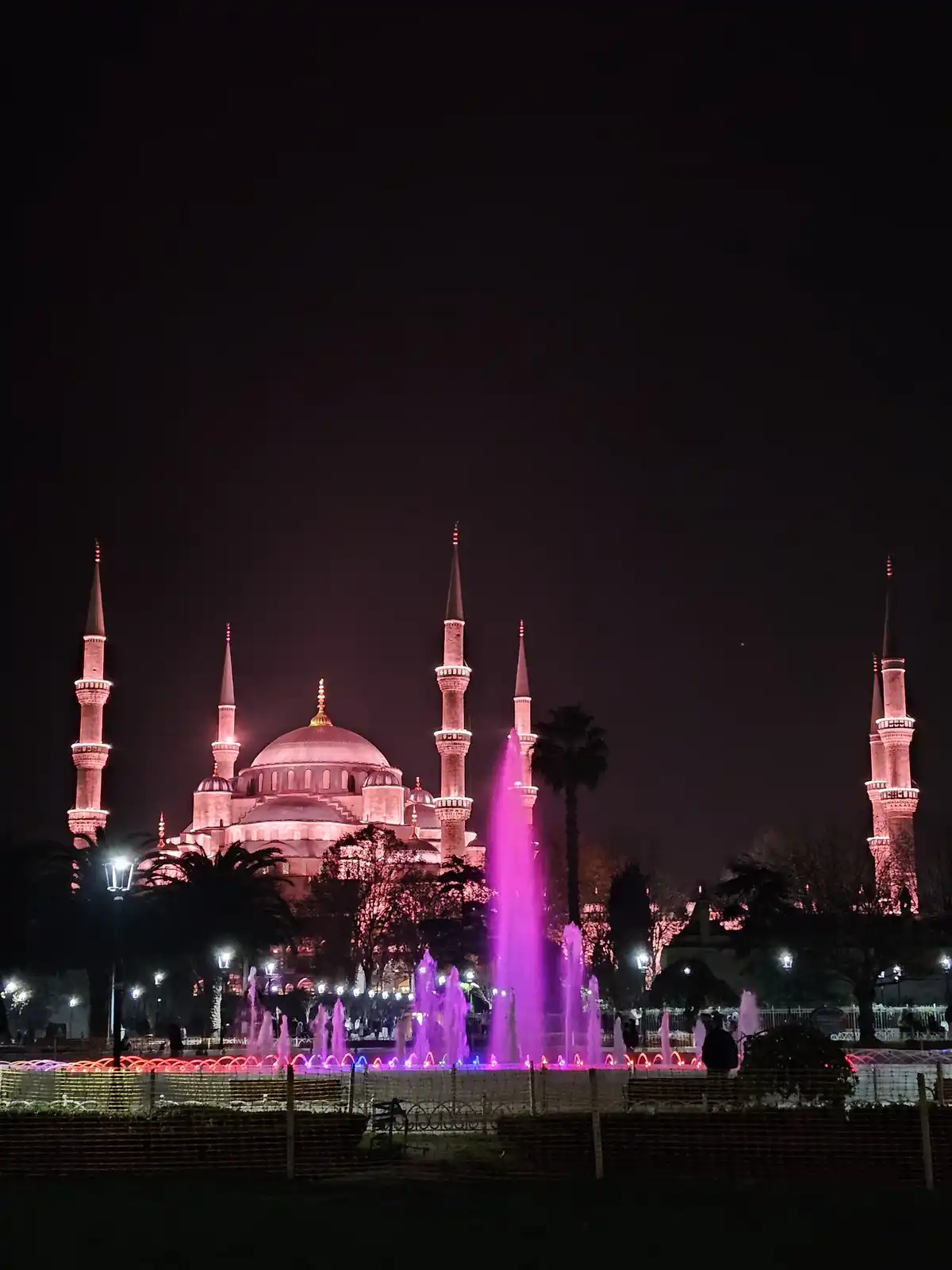 A nighttime photograph of the Blue Mosque in Istanbul, illuminated with pink and purple lights, with a vibrant…