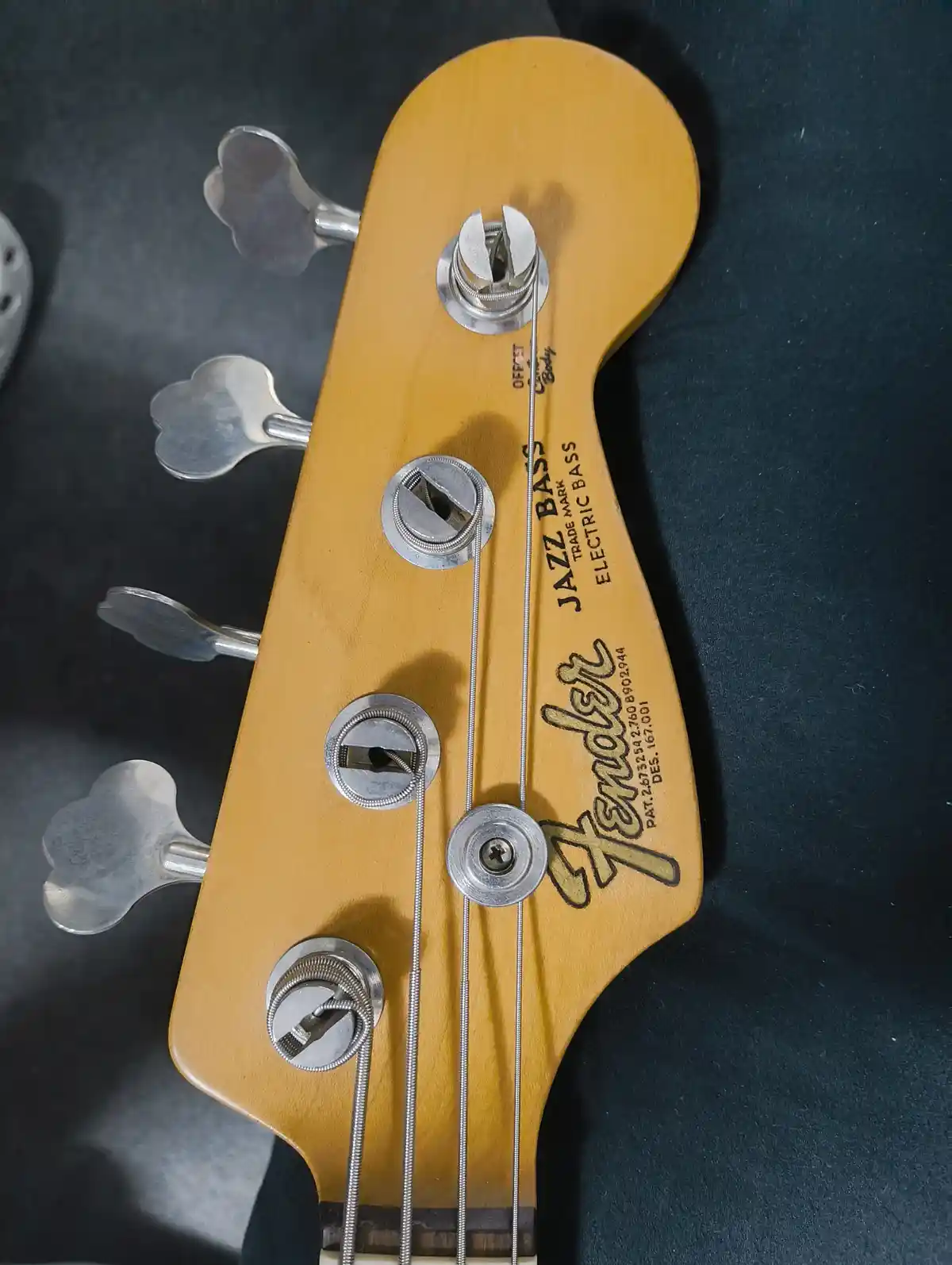 A close-up of a Fender Jazz Bass headstock with four silver tuning pegs and four strings running down the neck, showing the…