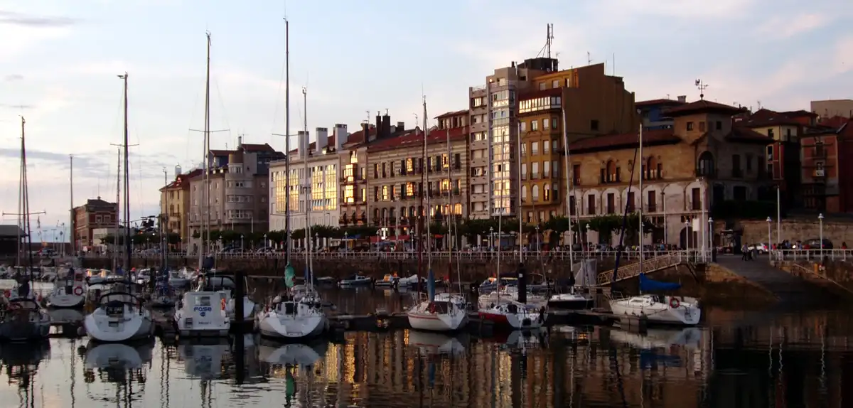A scenic harbor at dusk with sailboats docked in calm water, reflecting the warm glow of illuminated windows in multi-story…