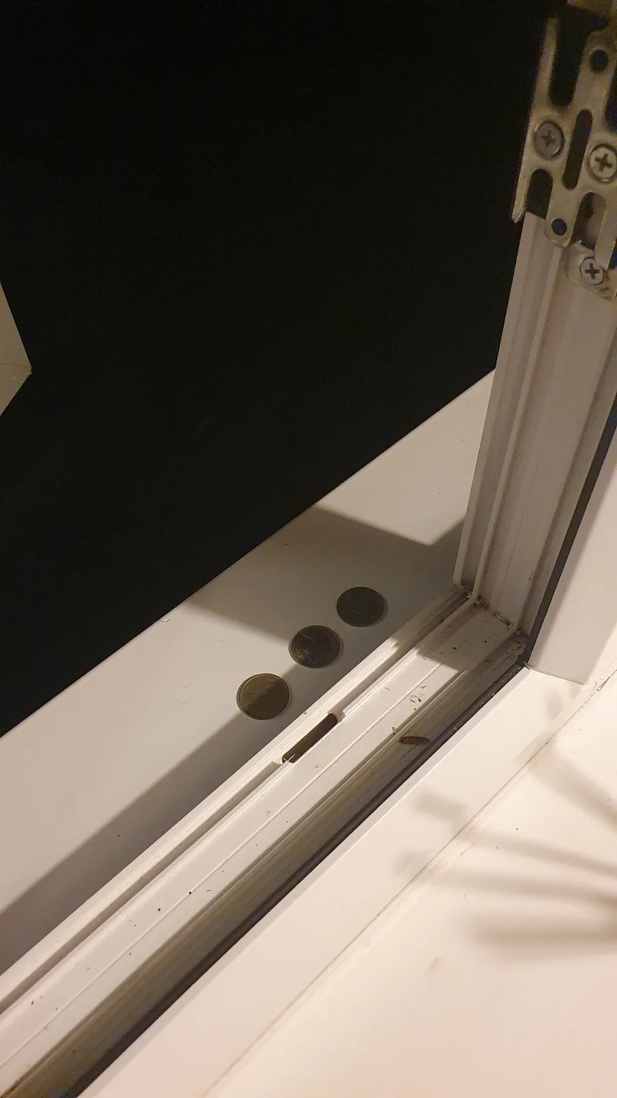 A close-up photo shows three coins lined up in the track of a sliding door's bottom seal, with a dark wall behind and a…