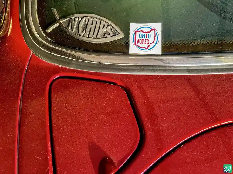 A close-up of a red car's window ledge shows a silver 'N'CHIPS' logo shaped like a fish and a white sticker with a red…