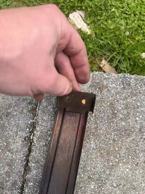 A hand holds a small metal bracket attached to a wooden fence rail, where the two holes in the bracket resemble eyes…