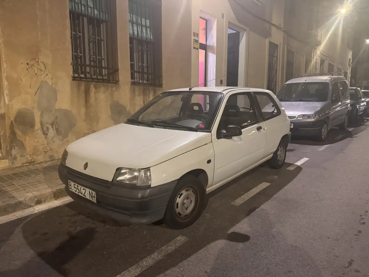 A white Renault 5 hatchback is parked on a narrow, dimly lit street at night, with a weathered building wall and other cars…