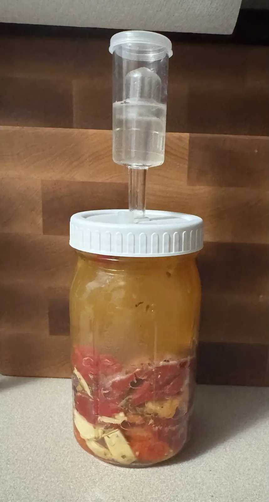 A glass mason jar filled with chopped red watermelon and pale yellow ginger pieces submerged in amber-colored liquid, topped…