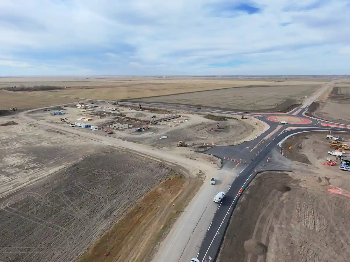 An aerial view shows a large, partially constructed road intersection with a newly paved roundabout in the center…