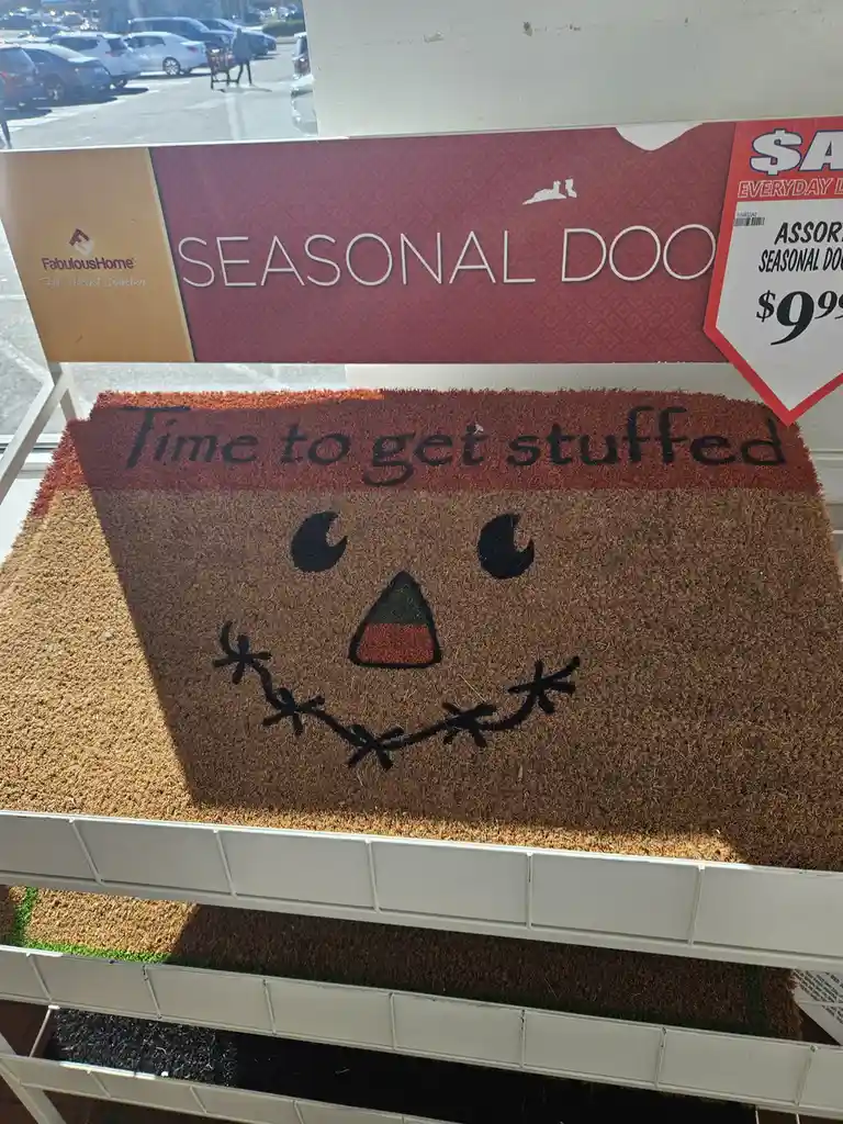 A brown coir doormat with a scarecrow face design, featuring crescent moon eyes, a candy corn nose, and stitched mouth, is…